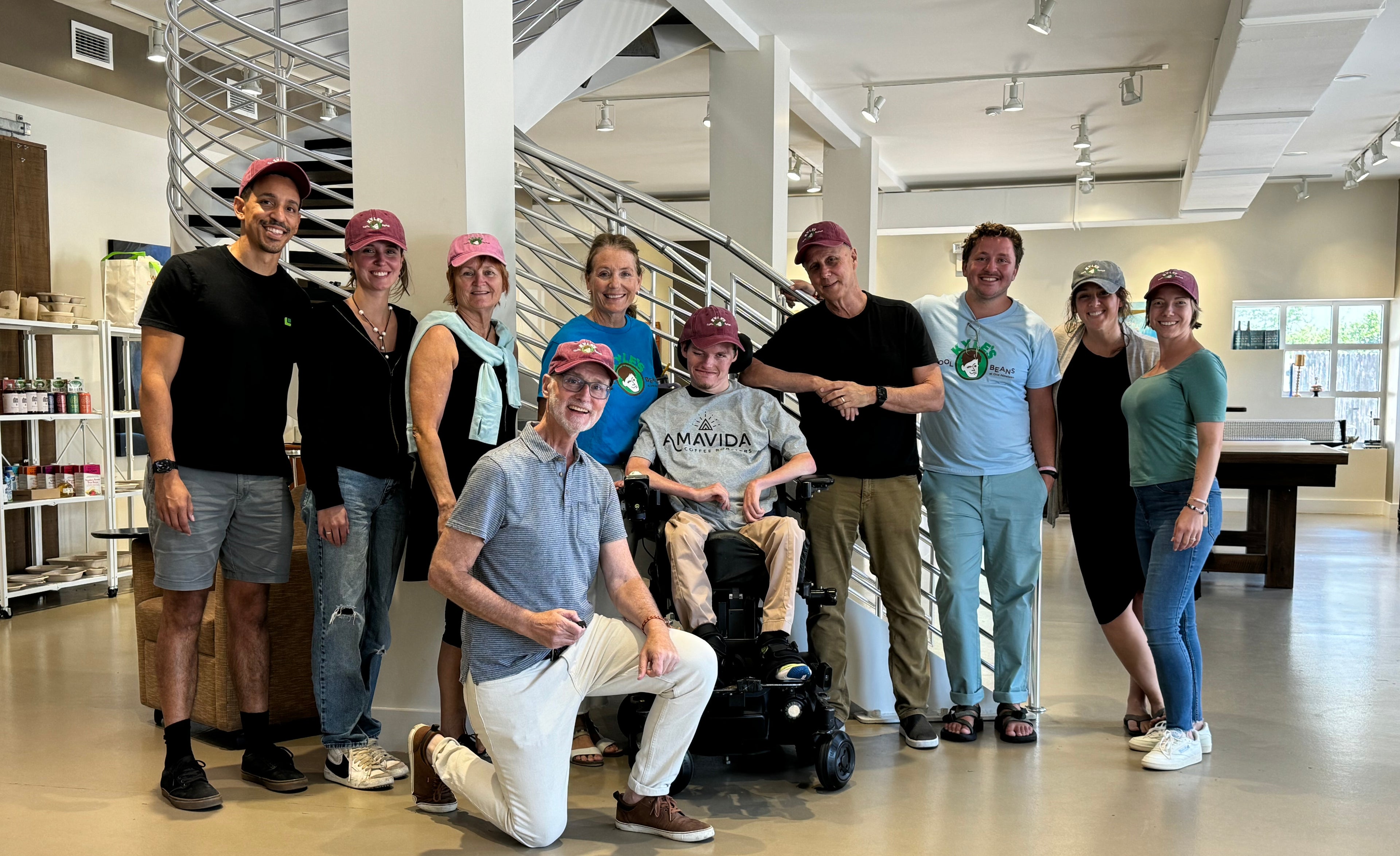 Kyle wearing an Amavida t-shirt sitting in his wheelchair. Kyle is surrounded by his family, friends, and Amavida employees. They are all smiling and most are wearing Kyle’s Cool Beans hats.