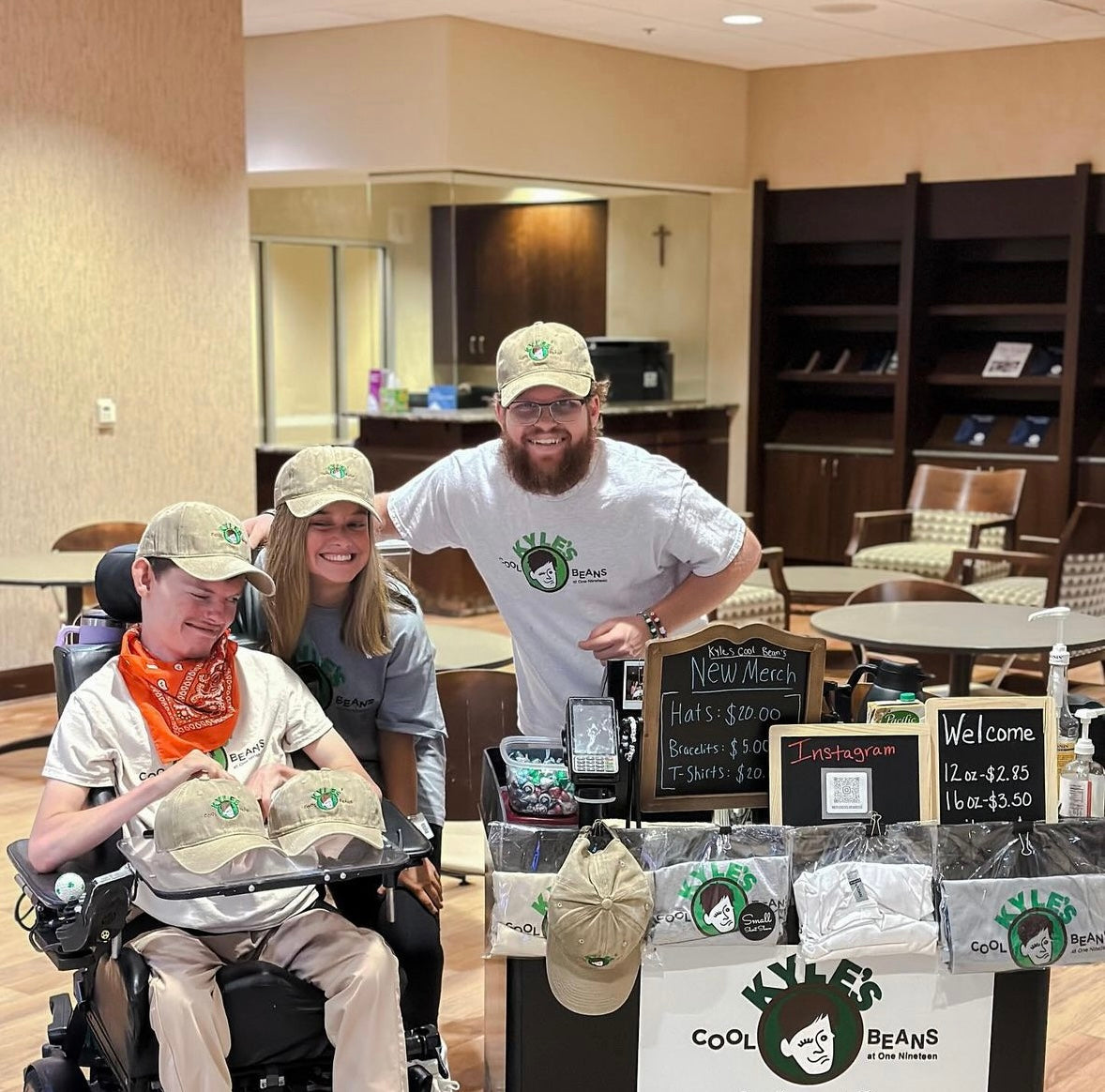 Kyle in his wheelchair next to his coffee cart, Lizzie and Jack are with him wearing Kyle’s Cool Beans T-shirts and hats!